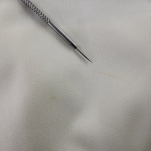 Puede incluir: Primer plano de una tela gris claro con un tejido texturizado. Una herramienta de metal plateado con un mango texturizado y una punta afilada se coloca diagonalmente sobre la tela. La herramienta parece ser una aguja o un instrumento similar.