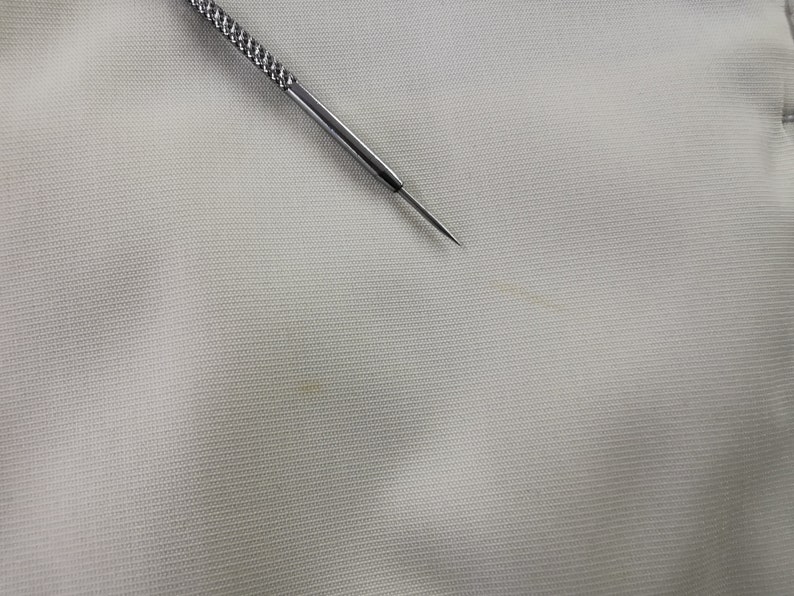 Puede incluir: Primer plano de una tela gris claro con un tejido texturizado. Una herramienta de metal plateado con un mango texturizado y una punta afilada se coloca diagonalmente sobre la tela. La herramienta parece ser una aguja o un instrumento similar.