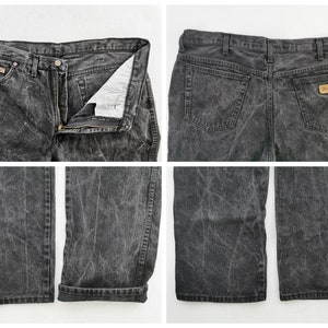 May include: A pair of acid-wash black denim jeans. The jeans have a button and zipper closure, front and back pockets, and a Wrangler brand patch. The jeans are shown from multiple angles, including front, back, and close-up views of the legs.