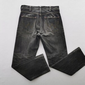 May include: Dark gray denim jeans with a faded, distressed appearance. Features classic five-pocket styling, a button and zipper closure, and the Wrangler logo on the back pockets. The jeans have a straight leg cut.