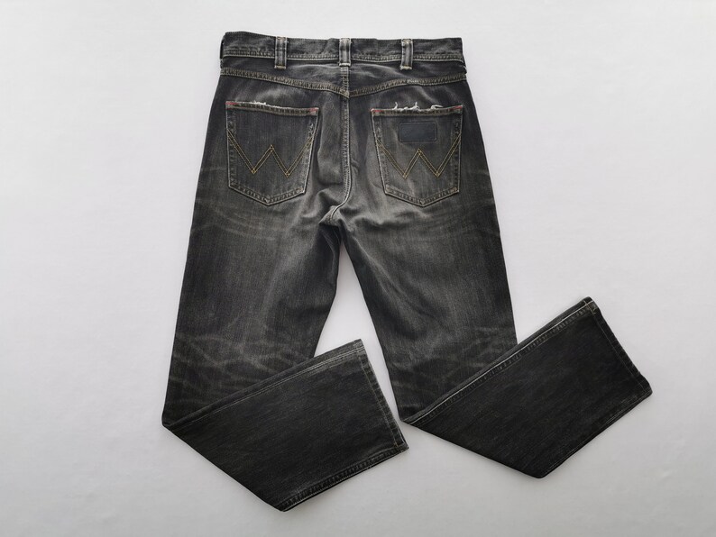 May include: Dark gray denim jeans with a faded, distressed appearance. Features classic five-pocket styling, a button and zipper closure, and the Wrangler logo on the back pockets. The jeans have a straight leg cut.