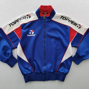 May include: A blue, red, and white track jacket with the brand name "TOPPER" printed on the sleeves and chest. The jacket has a full zipper and a ribbed collar and cuffs. The design features white panels on the shoulders and red accents.
