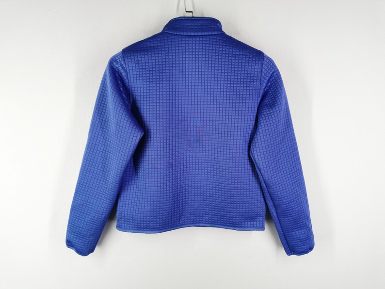May include: A blue zip-up jacket with a textured, grid-like pattern. The jacket has long sleeves and a stand-up collar. The garment is hanging against a white background.