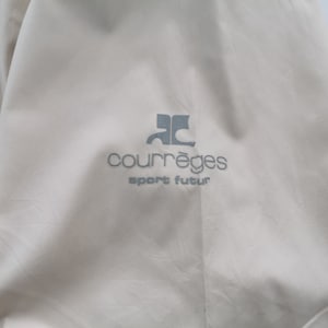 May include: A light beige garment featuring the grey embroidered logo of Courr&egrave;ges with the text sport futur positioned directly below the emblem. The fabric has a smooth, slightly matte texture with subtle folds visible across the surface area.