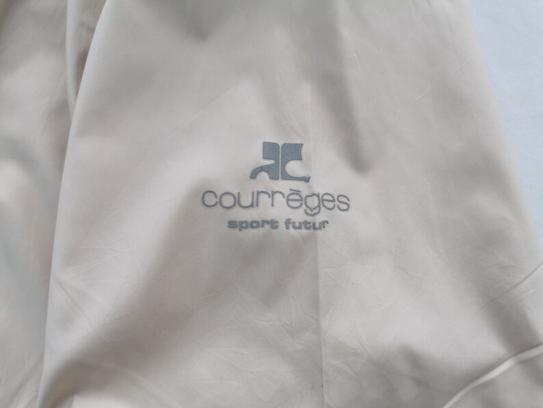May include: A light beige garment featuring the grey embroidered logo of Courr&egrave;ges with the text sport futur positioned directly below the emblem. The fabric has a smooth, slightly matte texture with subtle folds visible across the surface area.