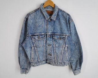 Pode incluir: Uma jaqueta jeans azul com lavagem ácida, gola clássica, mangas compridas e fecho frontal com botões. A peça inclui dois bolsos no peito com abas abotoadas e dois bolsos laterais. Uma etiqueta da marca Wrangler é visível.