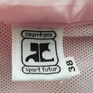 May include: A white fabric clothing label with black text reading courr&egrave;ges sport futur and the number 38, attached to a light pink garment with a white mesh lining. The label features a stylized black logo centered between the text lines.