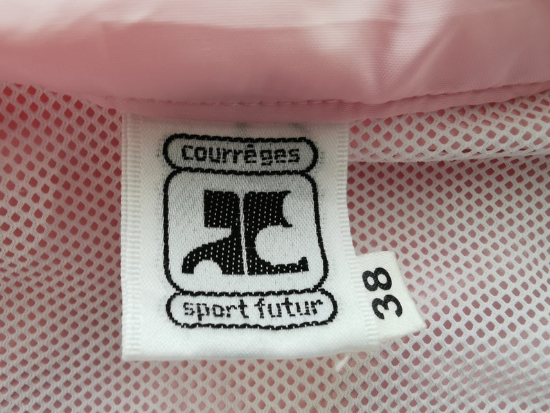 May include: A white fabric clothing label with black text reading courr&egrave;ges sport futur and the number 38, attached to a light pink garment with a white mesh lining. The label features a stylized black logo centered between the text lines.