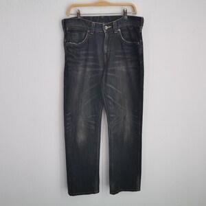 May include: Dark wash denim jeans hanging on a wooden hanger. The jeans have a classic five-pocket design, a button and zipper closure, and a faded, worn appearance. The jeans are straight-legged.