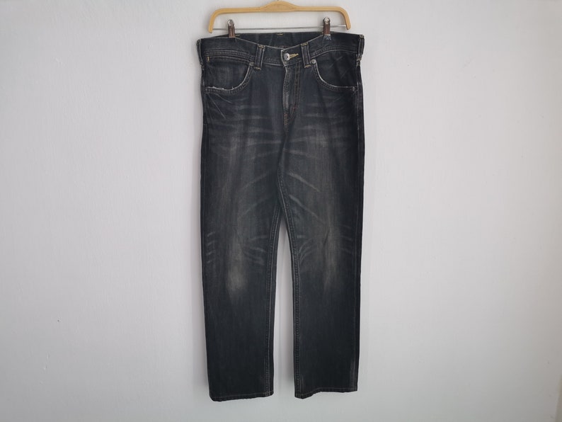 May include: Dark wash denim jeans hanging on a wooden hanger. The jeans have a classic five-pocket design, a button and zipper closure, and a faded, worn appearance. The jeans are straight-legged.