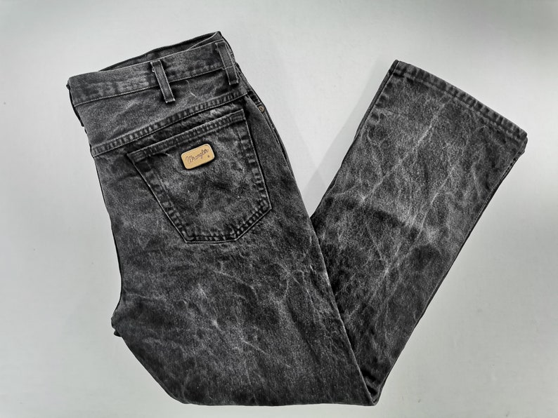 May include: Black acid wash Wrangler jeans. The jeans have a classic five-pocket design, a straight leg, and a faded appearance. The Wrangler logo is visible on the back pocket. A stylish and comfortable addition to any wardrobe.