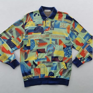 May include: A long-sleeved shirt with a colorful geometric pattern in shades of blue, yellow, red, and green. The shirt has a navy blue collar, cuffs, and hem. The text "COURR&Egrave;GES homme" is printed across the chest.