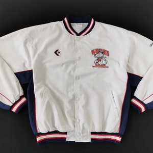 May include: A white snap-button jacket with navy blue and red accents. The front features a Converse logo on the right chest and a graphic of a wolf with the text "WOLVES" and "IHL-USA" on the left. It has striped ribbed cuffs, collar, and hem.