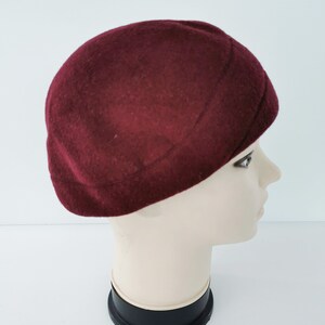 May include: A burgundy-colored beret hat. The hat is made of a soft, felt-like material and has a rounded crown. The beret is displayed on a mannequin head, showcasing its shape and style. The hat is a classic accessory.