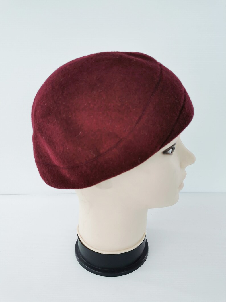 May include: A burgundy-colored beret hat. The hat is made of a soft, felt-like material and has a rounded crown. The beret is displayed on a mannequin head, showcasing its shape and style. The hat is a classic accessory.