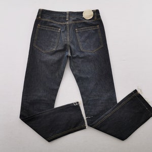 May include: Dark indigo denim jeans featuring a classic five-pocket design with contrast gold stitching. The pants have a straight-leg cut, a waistband with belt loops, and a circular beige paper tag attached to the back right side of the waist.