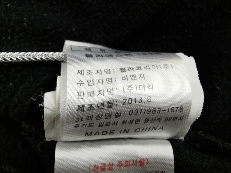 May include: A white tag with black text, attached to a black fabric. The tag includes text in Korean and English, with the words "MADE IN CHINA" at the bottom. The tag is attached with a silver string.