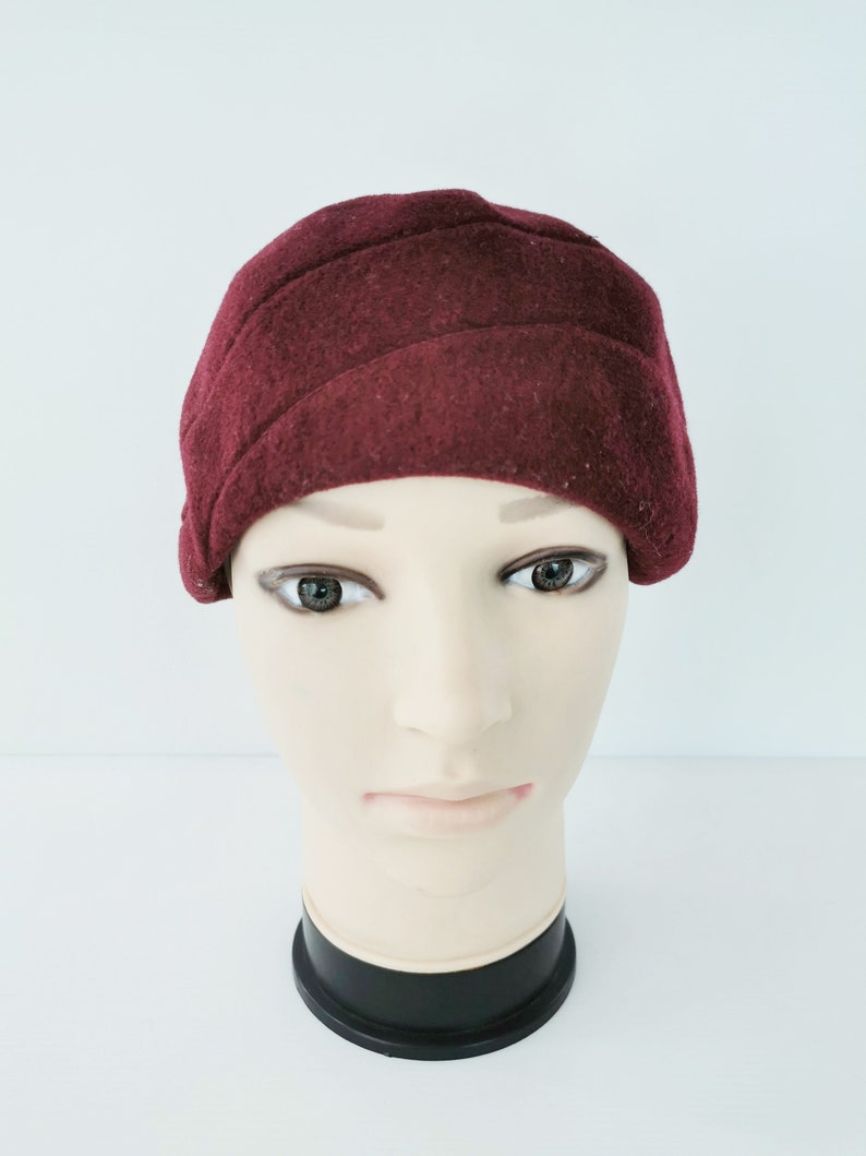 May include: A burgundy cloche hat with a ribbed design. The hat is made of a soft, felt-like material and is displayed on a mannequin head. The hat is a deep red color and has a classic, vintage style.