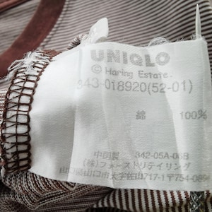 Pode incluir: Close-up de uma camiseta listrada marrom e branca com gola marrom. Uma etiqueta branca com a marca "UNIQLO" e outro texto est&aacute; presa &agrave; camisa. A etiqueta tamb&eacute;m indica que a camisa &eacute; 100% algod&atilde;o.