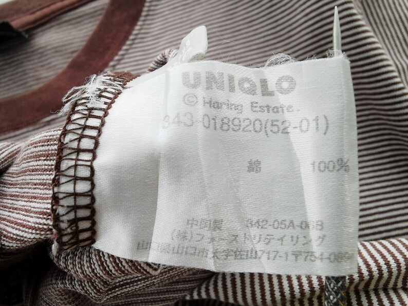 Pode incluir: Close-up de uma camiseta listrada marrom e branca com gola marrom. Uma etiqueta branca com a marca "UNIQLO" e outro texto est&aacute; presa &agrave; camisa. A etiqueta tamb&eacute;m indica que a camisa &eacute; 100% algod&atilde;o.