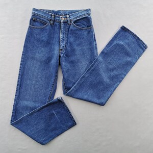 May include: A pair of classic blue denim jeans, laid flat against a neutral background. The jeans feature a button and zipper closure, front and back pockets, and belt loops. The denim has a medium wash and a straight leg cut.