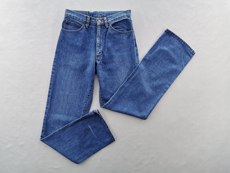 May include: A pair of classic blue denim jeans, laid flat against a neutral background. The jeans feature a button and zipper closure, front and back pockets, and belt loops. The denim has a medium wash and a straight leg cut.