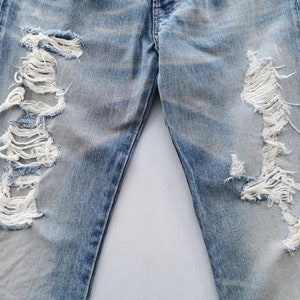 May include: Light blue denim jeans with distressed detailing. The jeans feature large, ripped sections on the front of the legs, revealing white threads. The denim has a faded appearance.
