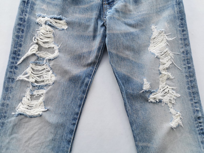 May include: Light blue denim jeans with distressed detailing. The jeans feature large, ripped sections on the front of the legs, revealing white threads. The denim has a faded appearance.