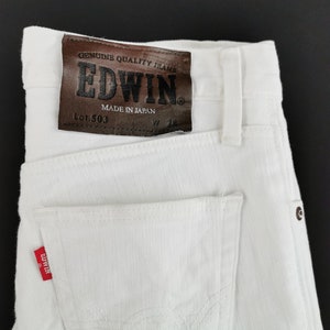 K&ouml;nnte beinhalten: Nahaufnahme einer wei&szlig;en Jeans aus Denim mit einem braunen Lederaufn&auml;her. Der Aufn&auml;her tr&auml;gt die Aufschrift "EDWIN" und "MADE IN JAPAN". Die Jeans hat ein rotes "EDWIN"-Etikett auf der Ges&auml;&szlig;tasche.
