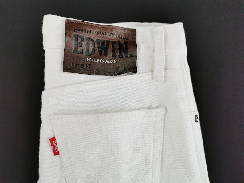K&ouml;nnte beinhalten: Nahaufnahme einer wei&szlig;en Jeans aus Denim mit einem braunen Lederaufn&auml;her. Der Aufn&auml;her tr&auml;gt die Aufschrift "EDWIN" und "MADE IN JAPAN". Die Jeans hat ein rotes "EDWIN"-Etikett auf der Ges&auml;&szlig;tasche.
