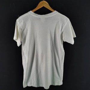 May include: A plain white t-shirt hanging on a wooden hanger against a dark background. The short-sleeved shirt is made of a soft, lightweight fabric. The back of the shirt is visible, showing the neckline and the shoulders.