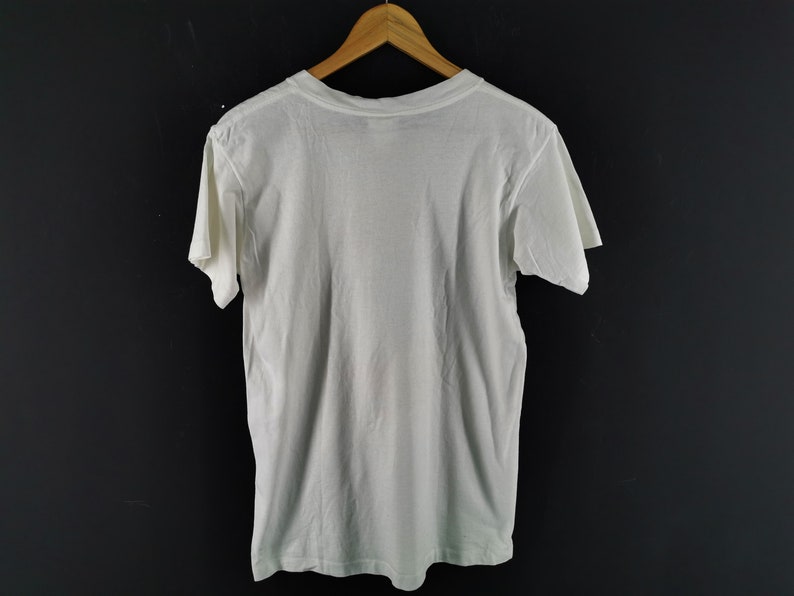 May include: A plain white t-shirt hanging on a wooden hanger against a dark background. The short-sleeved shirt is made of a soft, lightweight fabric. The back of the shirt is visible, showing the neckline and the shoulders.