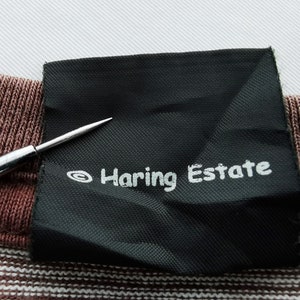 Pode incluir: Close-up de uma etiqueta preta com o texto "Haring Estate" em branco. A etiqueta est&aacute; em um tecido listrado marrom e branco. Uma agulha prateada aponta para a etiqueta.