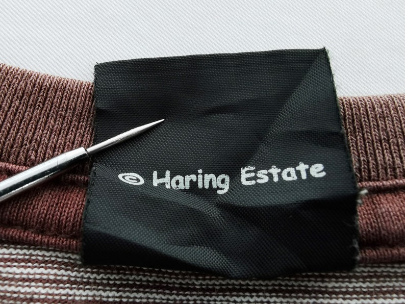 Pode incluir: Close-up de uma etiqueta preta com o texto "Haring Estate" em branco. A etiqueta est&aacute; em um tecido listrado marrom e branco. Uma agulha prateada aponta para a etiqueta.