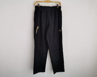 Puede incluir: Pantalones deportivos negros con cintura elástica y perneras rectas. Los pantalones muestran un logotipo dorado de Mizuno en el muslo izquierdo y el texto vertical dorado de Mizuno en el lado derecho. La prenda está colgada de una percha de madera.