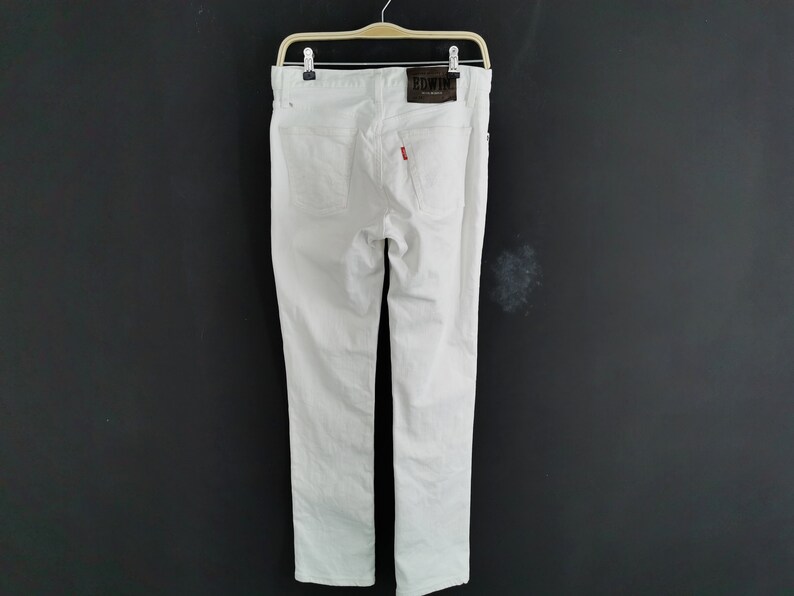 K&ouml;nnte beinhalten: Wei&szlig;e Jeans aus Denim, die an einem Holzb&uuml;gel h&auml;ngen. Die Jeans haben ein klassisches F&uuml;nf-Taschen-Design, ein rotes Logo-Etikett und einen braunen Lederaufn&auml;her mit dem Markennamen. Die Jeans sind ein sauberes, knackiges Wei&szlig;.