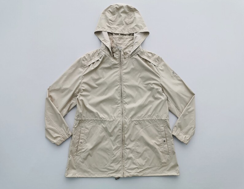 May include: A beige hooded windbreaker jacket featuring a full-length front zipper, long sleeves with elasticized cuffs, and two front pockets with snap button closures. The jacket includes a drawstring waist and shoulder epaulet details.