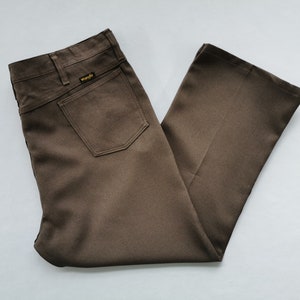 Peut inclure: Un pantalon marron uni de la marque Wrangler avec une taille haute, une poche arrière unique ornée d'une petite étiquette jaune et noire, des passants de ceinture et une coupe droite. Le tissu présente une texture lisse et uniforme.