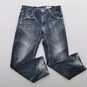 May include: Dark wash denim jeans with a faded effect. The jeans have a classic five-pocket design, a button and zip closure, and distressed details at the hem. The jeans are laid flat on a white surface.