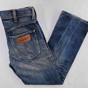 Może przedstawiać: Niebieskie dżinsy z klasycznym pięciokieszeniowym designem. Dżinsy mają wyblakłe pranie i logo "Wrangler" na tylnej kieszeni. Dżinsy mają prosty kr&oacute;j nogawki.
