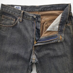 May include: Close-up of dark grey denim jeans with an unzipped fly. The jeans feature a classic five-pocket design, a button closure, and a small label reading "EDWIN". The interior lining is a contrasting brown colour.