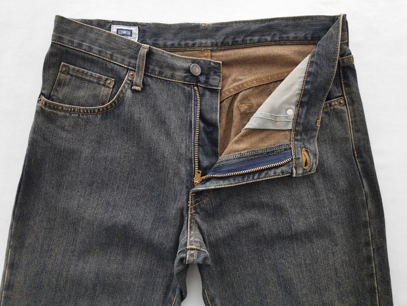 May include: Close-up of dark grey denim jeans with an unzipped fly. The jeans feature a classic five-pocket design, a button closure, and a small label reading "EDWIN". The interior lining is a contrasting brown colour.