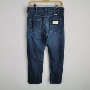 May include: Dark blue denim jeans hanging on a wooden hanger. The jeans have a classic five-pocket design, with the Wrangler logo visible on the back pocket. The jeans are a medium wash and appear to be unworn.