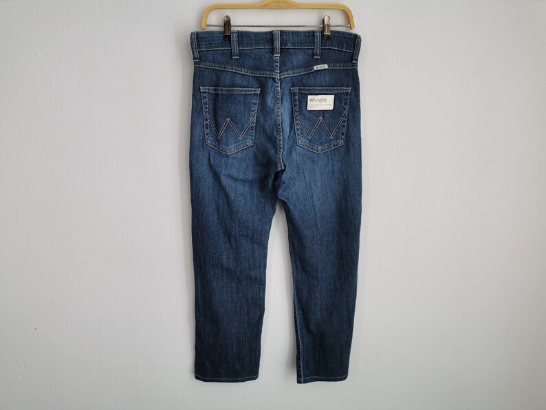 May include: Dark blue denim jeans hanging on a wooden hanger. The jeans have a classic five-pocket design, with the Wrangler logo visible on the back pocket. The jeans are a medium wash and appear to be unworn.