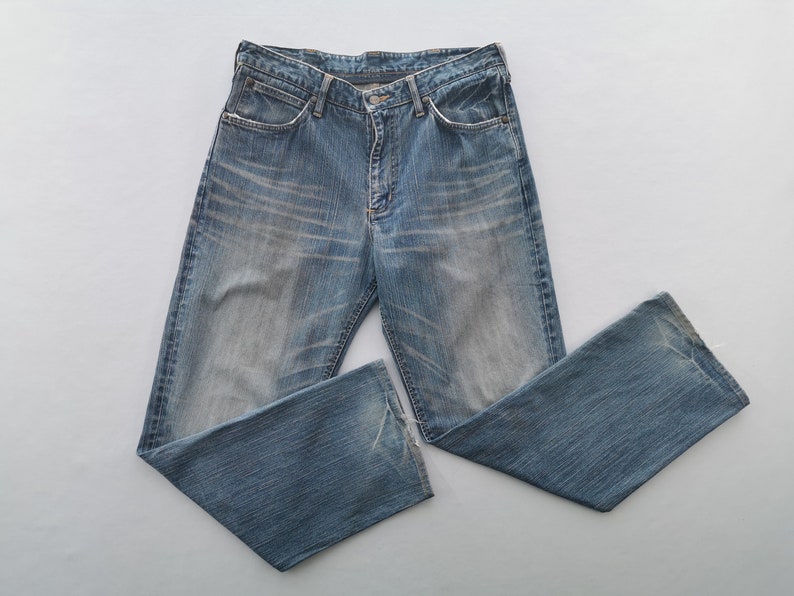 May include: Light blue denim jeans with a faded wash and a classic five-pocket design. The jeans have a button and zipper closure and are laid flat on a white surface. The jeans are a standard fit.