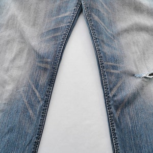 May include: Close-up of light blue denim jeans with distressed detailing. The jeans have a faded wash and a ripped section on the right leg. The seams are visible, and the fabric shows wear.
