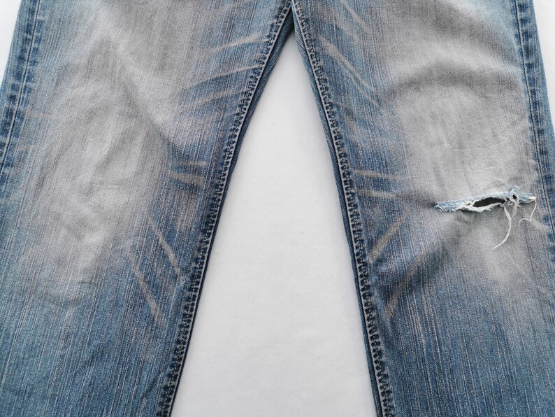 May include: Close-up of light blue denim jeans with distressed detailing. The jeans have a faded wash and a ripped section on the right leg. The seams are visible, and the fabric shows wear.