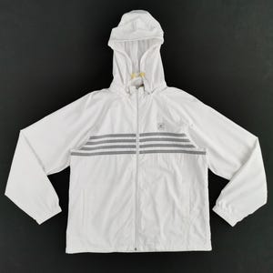 Puede incluir: Chaqueta blanca con capucha, cierre de cremallera y rayas horizontales grises en el pecho. La chaqueta tiene un peque&ntilde;o logotipo y una capucha con cord&oacute;n. Las mangas son largas y los pu&ntilde;os el&aacute;sticos.