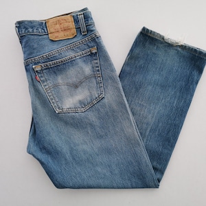 May include: Blue denim jeans featuring a faded wash, a tan leather patch on the back waistband, a single back pocket with a small red tab, and frayed edges at the bottom hem. The jeans have a classic five-pocket construction with metal rivets.