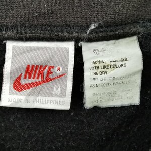 K&ouml;nnte beinhalten: Nahaufnahme eines schwarzen Kleidungsst&uuml;cks mit zwei Etiketten. Das linke Etikett zeigt das rote Nike-Logo und das Wort "NIKE" in Wei&szlig;. Das rechte Etikett enth&auml;lt Waschanweisungen. Das Kleidungsst&uuml;ck wird in den Philippinen hergestellt.